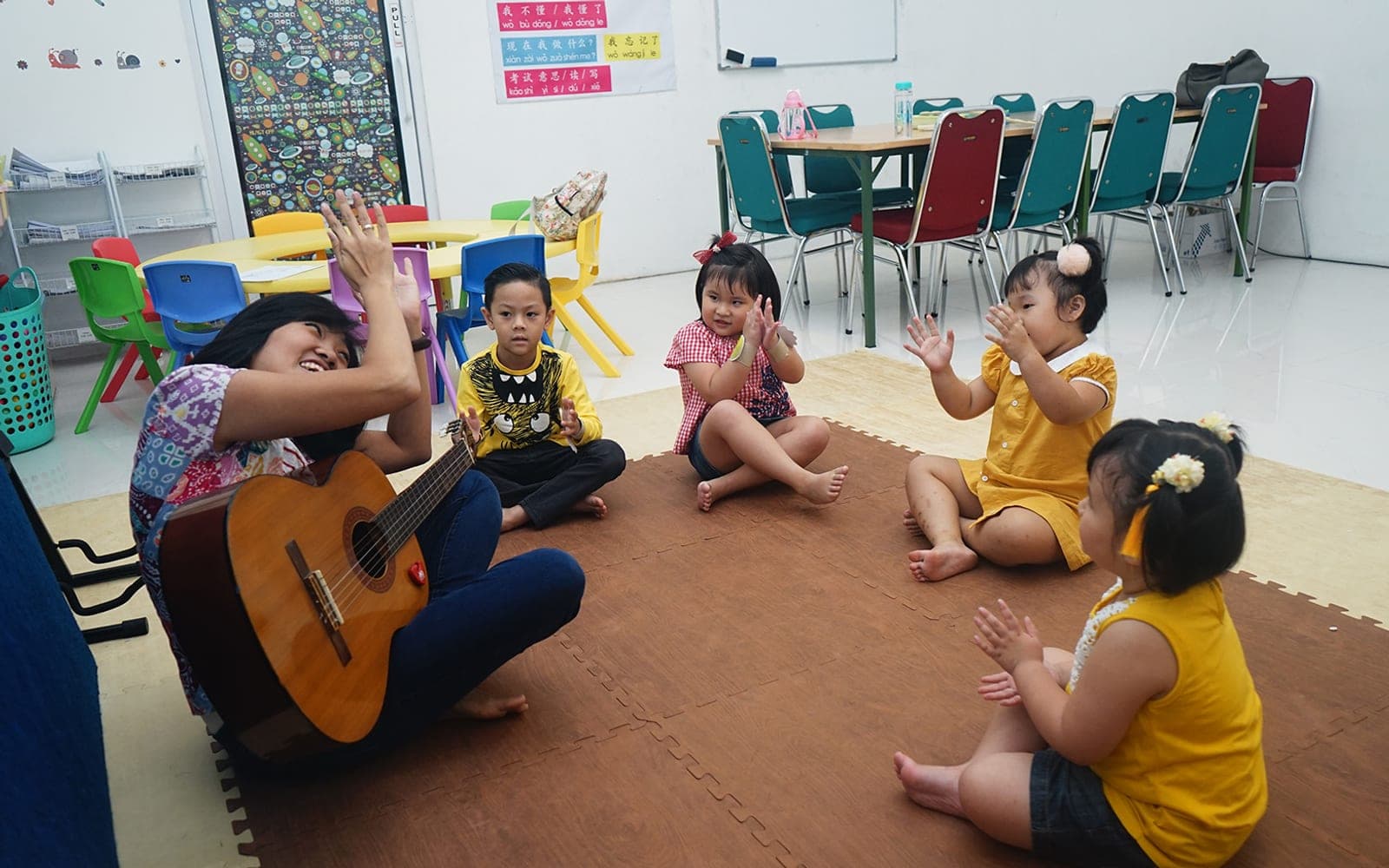 Guru Kiddles bermain gitar sambil menyanyi bersama anak-anak kelas Kinder yang duduk melingkar di lantai